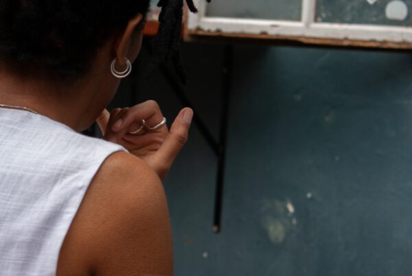 partial back view of person of colour with their hand lifted to their mouth with a blue coloured wall in the background